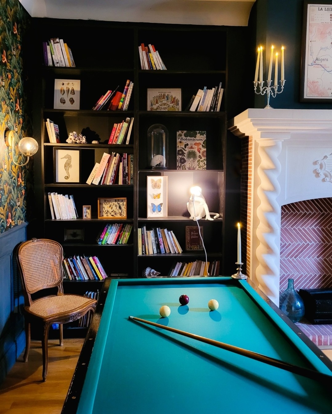 A billiard table is positioned in the foreground, accompanied by a cue stick and colorful balls. In the background, a dark bookshelf filled with various books and decorative items is visible. A chair rests nearby, while soft candlelight enhances the cozy ambiance created by the fireplace.