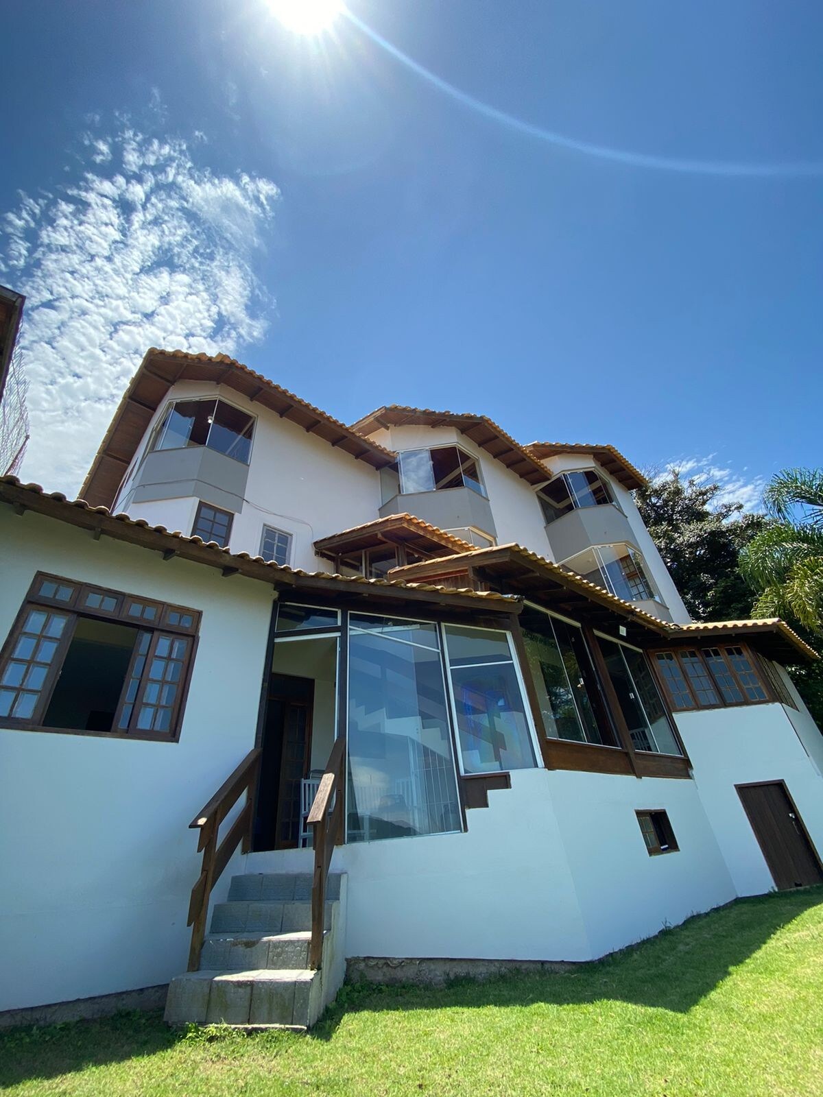 The exterior of a multi-story house is presented, featuring a combination of wooden accents and large windows that allow natural light to fill the spaces. A staircase leads to the entrance, with a lush green lawn visible in the foreground. Bright blue skies add to the overall aesthetic.