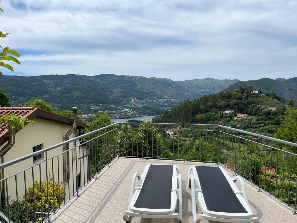 A terrace features two lounge chairs positioned to provide unobstructed views of the surrounding hills and the river below. The landscape showcases a variety of green hues, complemented by a clear sky above. The house is partially visible to the left.