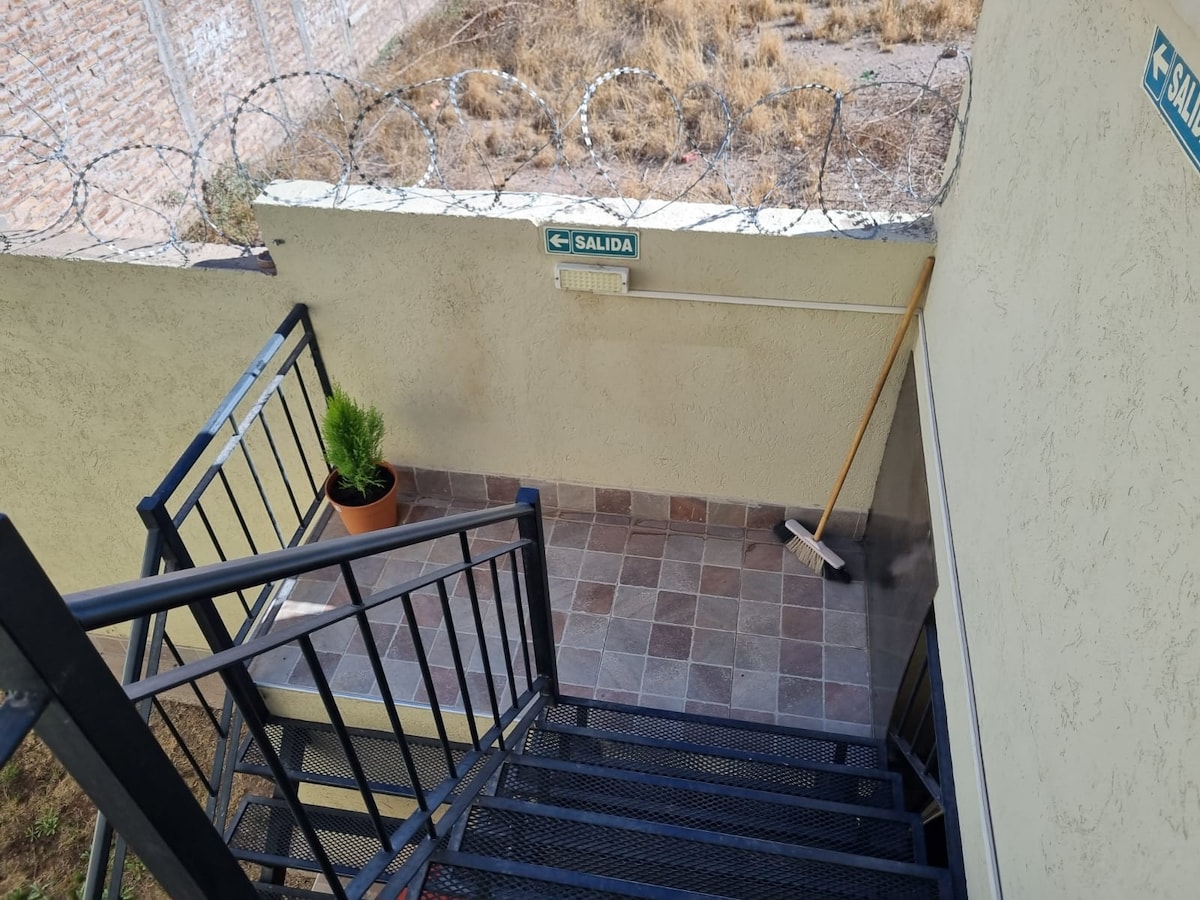 A stairway leads to a well-maintained exit area. The space is adorned with a small potted plant, and a broom is positioned against the wall. The tiled floor features a neutral color palette, and a barbed wire fence is visible above.