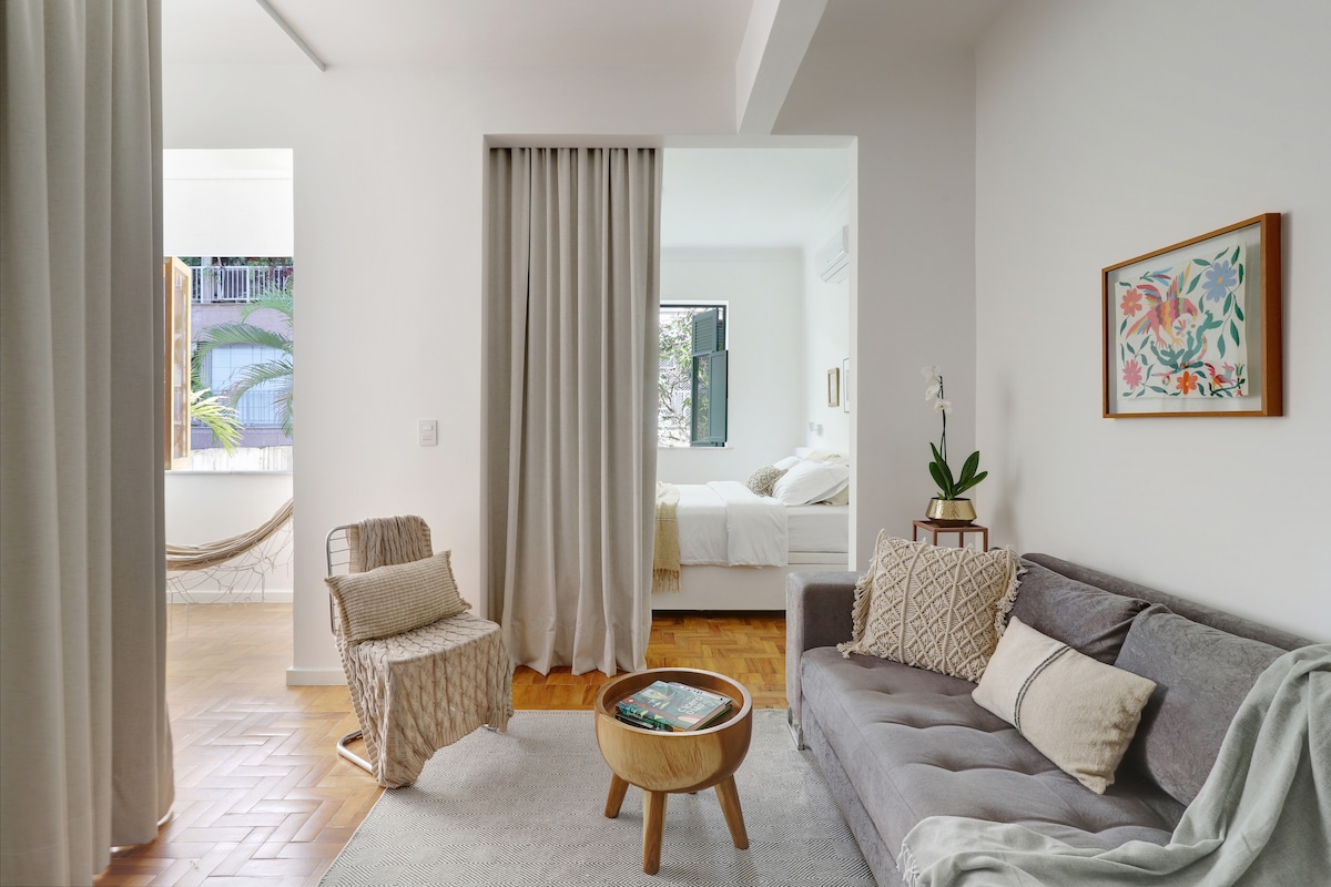 A cozy living area features a soft gray sofa with decorative pillows and a round coffee table. Curtains frame an entrance to the sleeping area, where natural light brightens a spacious bed. A small plant and framed artwork add warmth to the room.