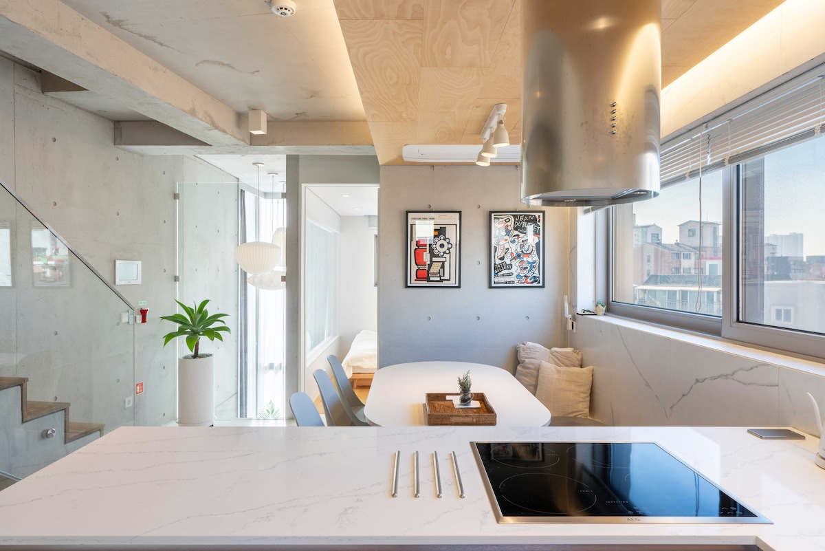A modern kitchen space features a large white countertop, a sleek stovetop, and ample natural light from the windows. Framed artwork adorns the walls, and a small indoor plant adds a touch of greenery. The stylish staircase is visible in the background.