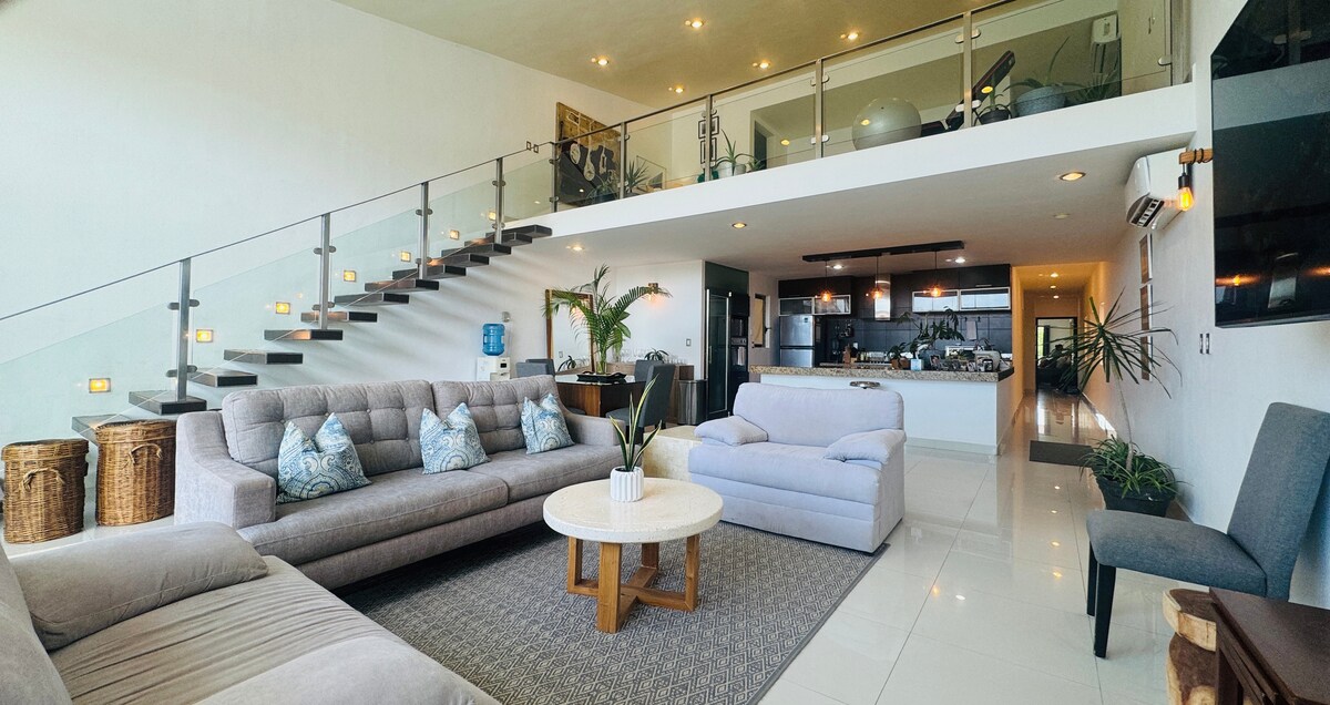 The open-concept living area features a comfortable seating arrangement of gray sofas and accent pillows. A round wooden coffee table sits atop a textured rug. The dramatic staircase, with glass railings, leads to an upper level, while a modern kitchen serves as the backdrop.