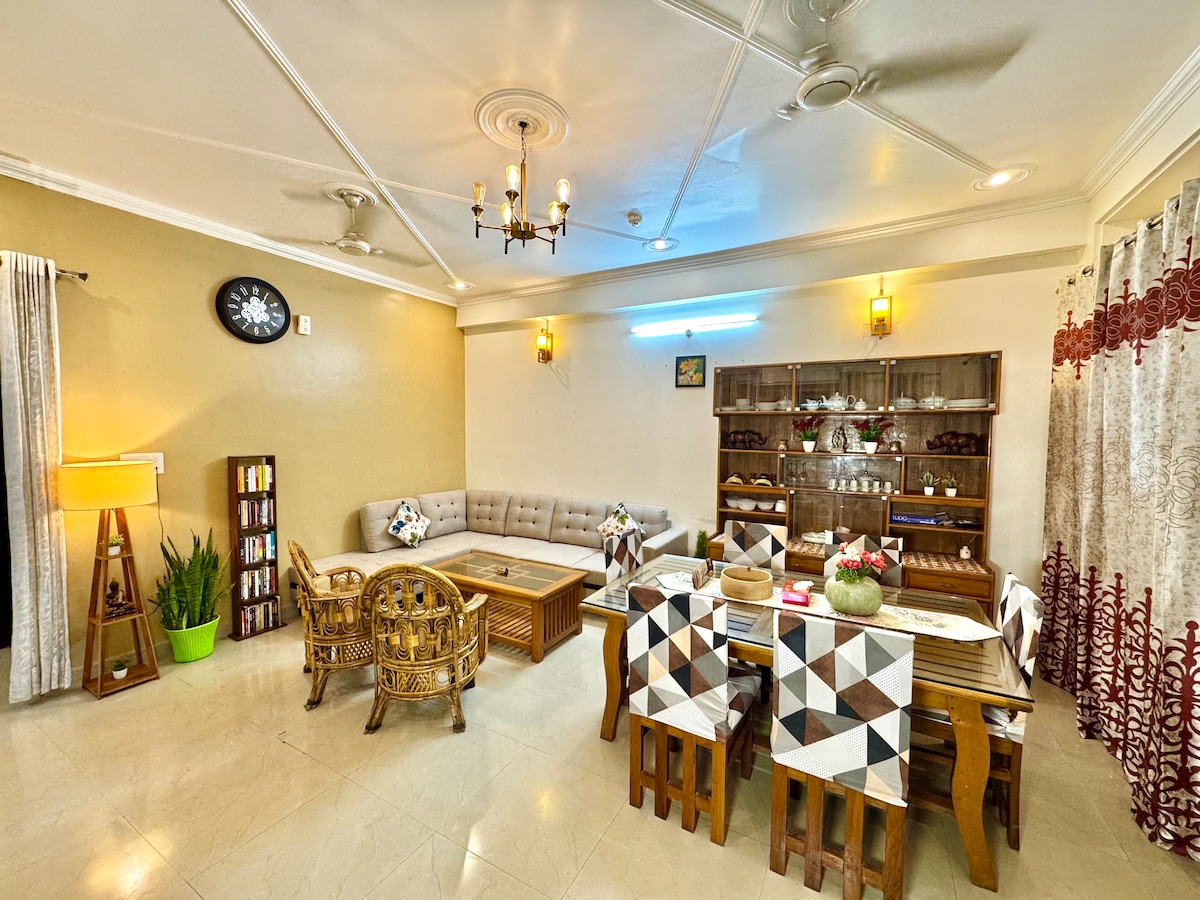 A spacious living area features a large sectional sofa in neutral tones, complemented by a wooden coffee table. A dining set with patterned chair covers is placed nearby. Decorative plants and soft lighting enhance the inviting ambiance, while curtains frame the windows, allowing natural light to fill the room.