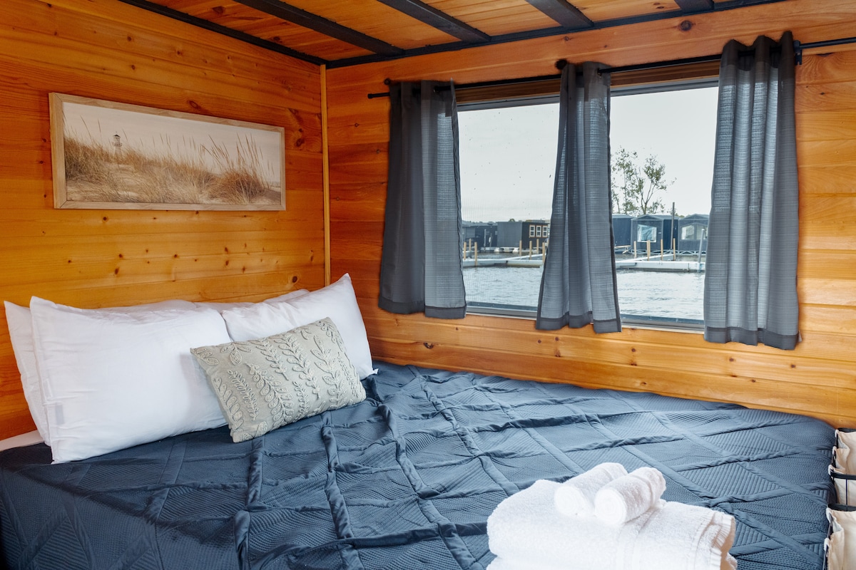 A snug sleeping area features warm wooden paneling and large windows offering views of the surrounding marina. A spacious bed is adorned with crisp white pillows and a decorative throw pillow. Rolled towels are placed neatly on the bed, enhancing the inviting appearance.