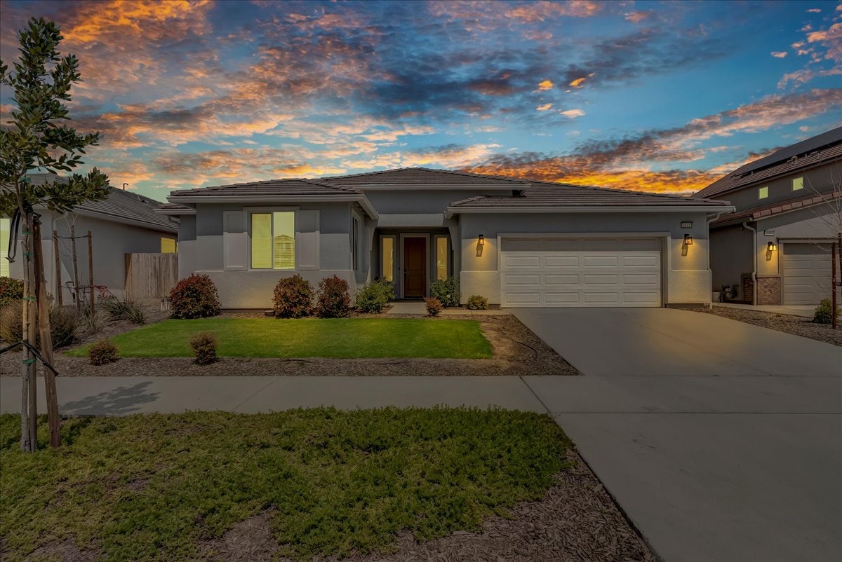 The exterior of a modern home is presented, showcasing a well-maintained yard with shrubs and a driveway leading to a three-car garage. The entrance is framed by subtle outdoor lighting, and the vibrant sky above features striking colors at dusk.