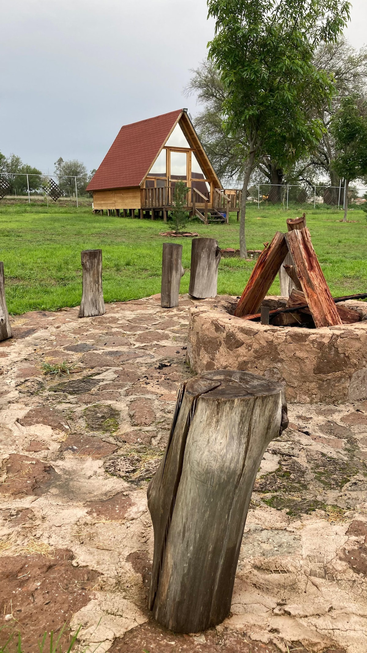 A rustic stone fire pit is surrounded by tree stump seating, with an A-frame cabin visible in the background. Lush green grass expands throughout the area, complemented by scattered trees and an overcast sky.