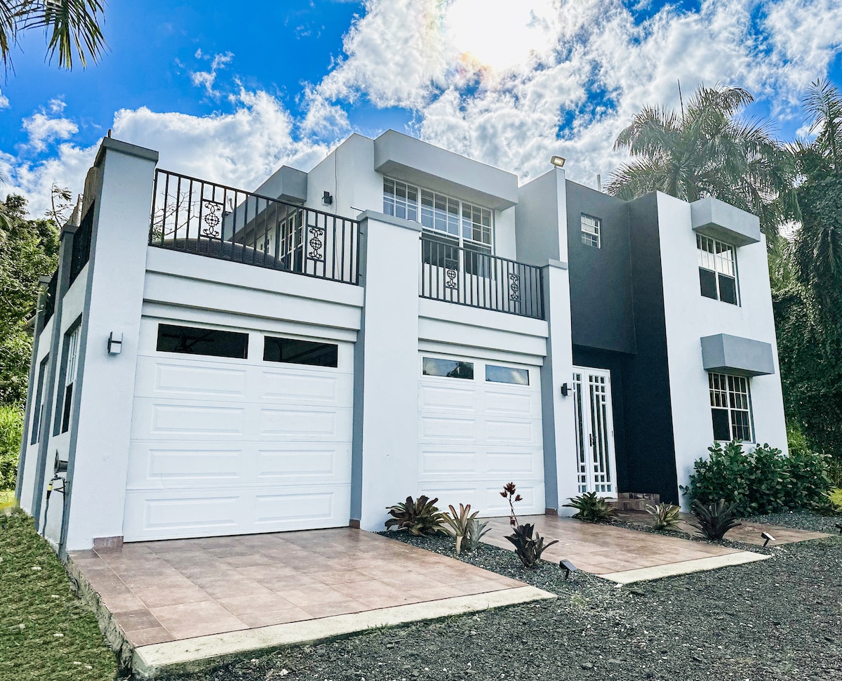 The exterior of the home is presented, featuring a modern two-story design with a combination of white and dark accents. Two garage doors are visible, along with a stylish entrance lit by natural light. Lush greenery and tropical plants frame the entrance.