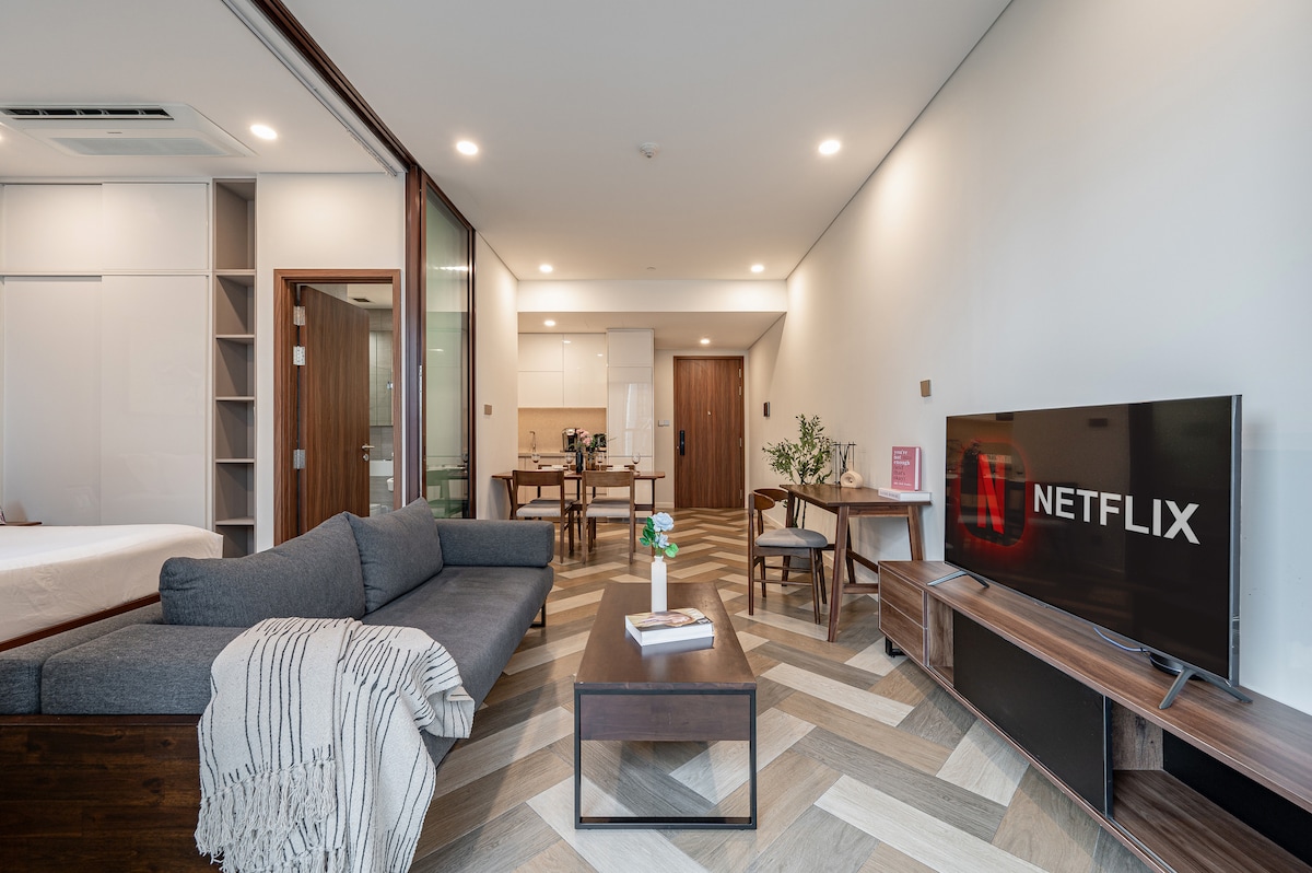A modern living area features a comfortable sofa facing a 55-inch TV with Netflix. A dining table and chairs are visible in the background, along with a fully equipped kitchen. Natural light enters through large glass doors, enhancing the spaciousness.