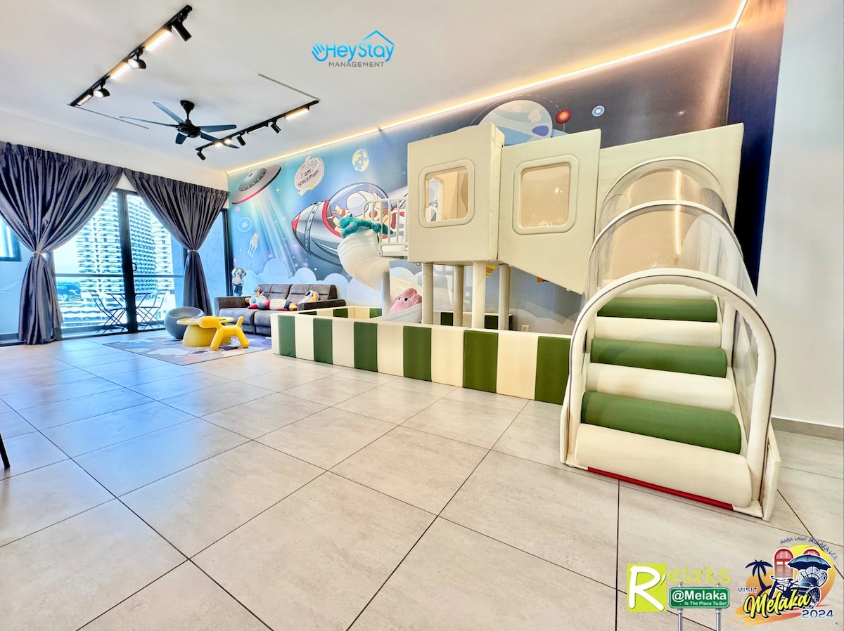 A colorful play area features a soft slide and play structure against a whimsical mural. Large windows allow natural light, highlighting the spacious floor design. Brightly colored furniture pieces are scattered throughout, creating an inviting environment for families.