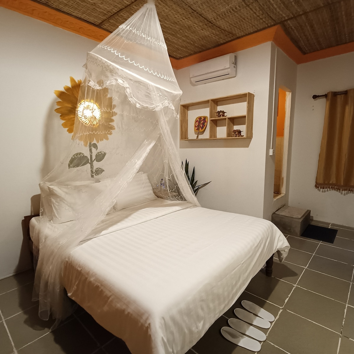 A cozy sleeping area features a spacious bed draped with a delicate mosquito net, creating a serene atmosphere. A sunflower mural adds a touch of brightness to the wall, while a soft light fixture illuminates the space. Slippers are neatly placed on the floor beside the bed.