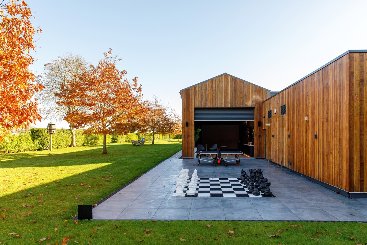 An outdoor area showcases a large chessboard set within a manicured lawn, surrounded by trees displaying autumn foliage. The space features a modern wooden structure with a sleek façade, creating a serene environment for relaxation and outdoor activities.
