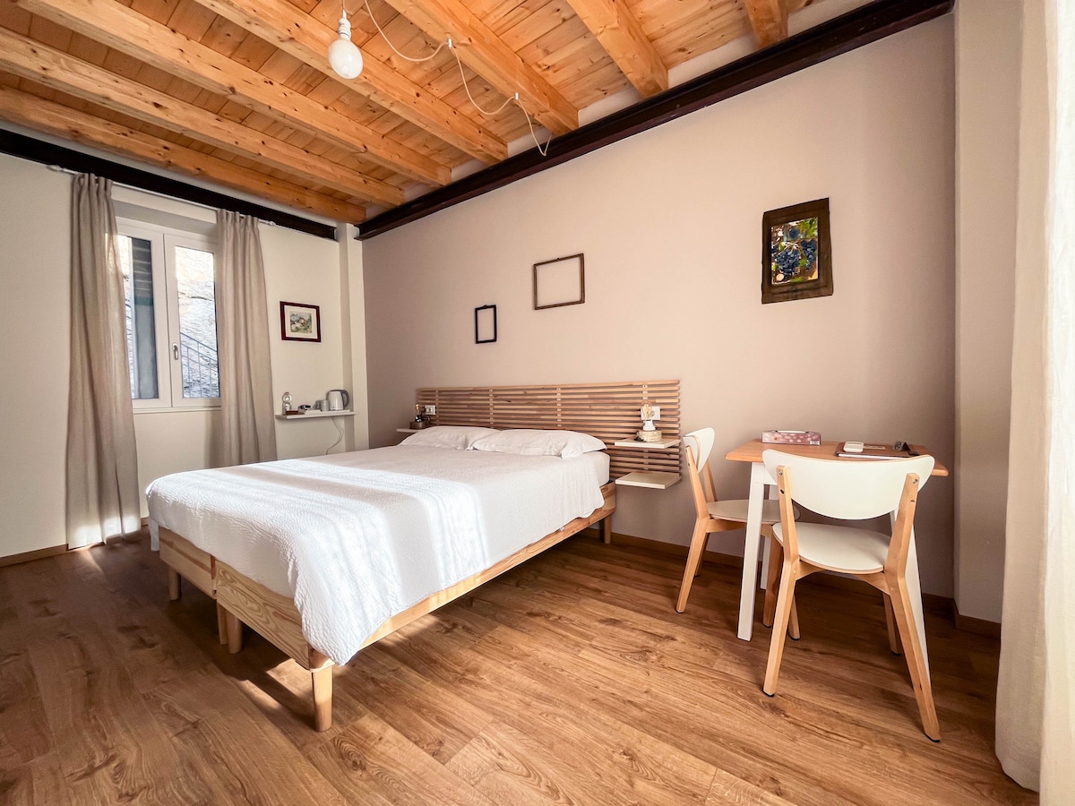A well-lit room features a spacious bed against a light-toned wall. Wooden beams run across the ceiling, adding warmth. A small desk with a chair is positioned near a window, which is dressed with soft curtains. Artwork decorates the walls, enhancing the inviting space.