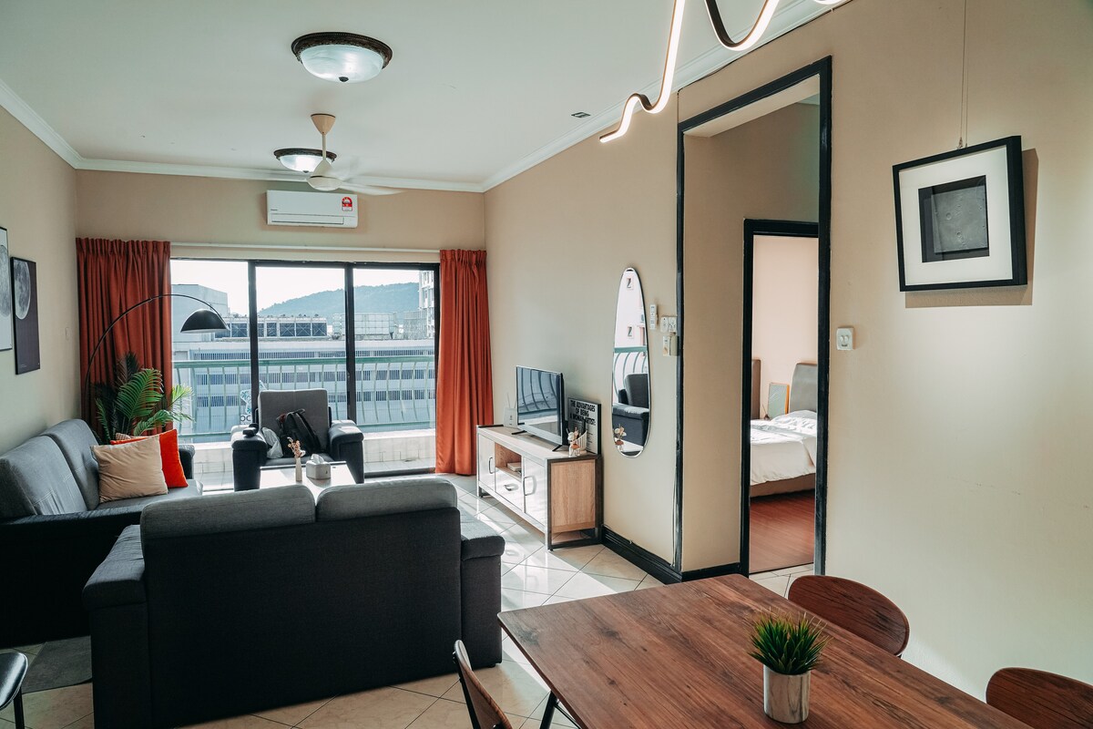 A spacious living area is presented with large windows offering natural light. Two comfortable sofas face a small television, while a dining table with four chairs is visible in the foreground. Bright curtains frame the windows, and a mirror is positioned near the entrance.