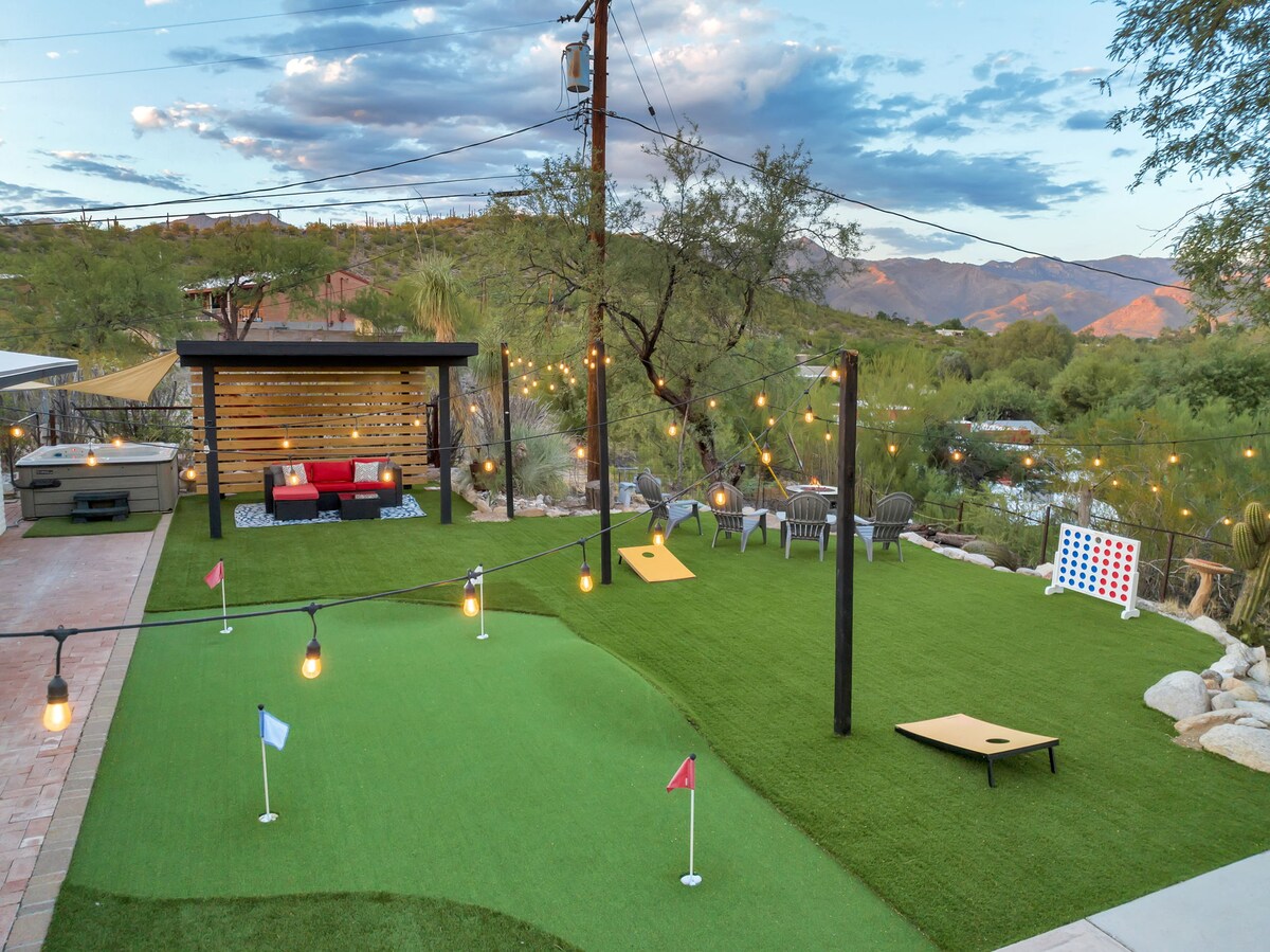 An outdoor space features a putting green with four holes and vibrant mountain views in the background. Cozy seating arrangements include a red sofa and several lounge chairs. Soft lighting is provided by hanging bulbs, creating an inviting atmosphere for relaxation and entertainment.