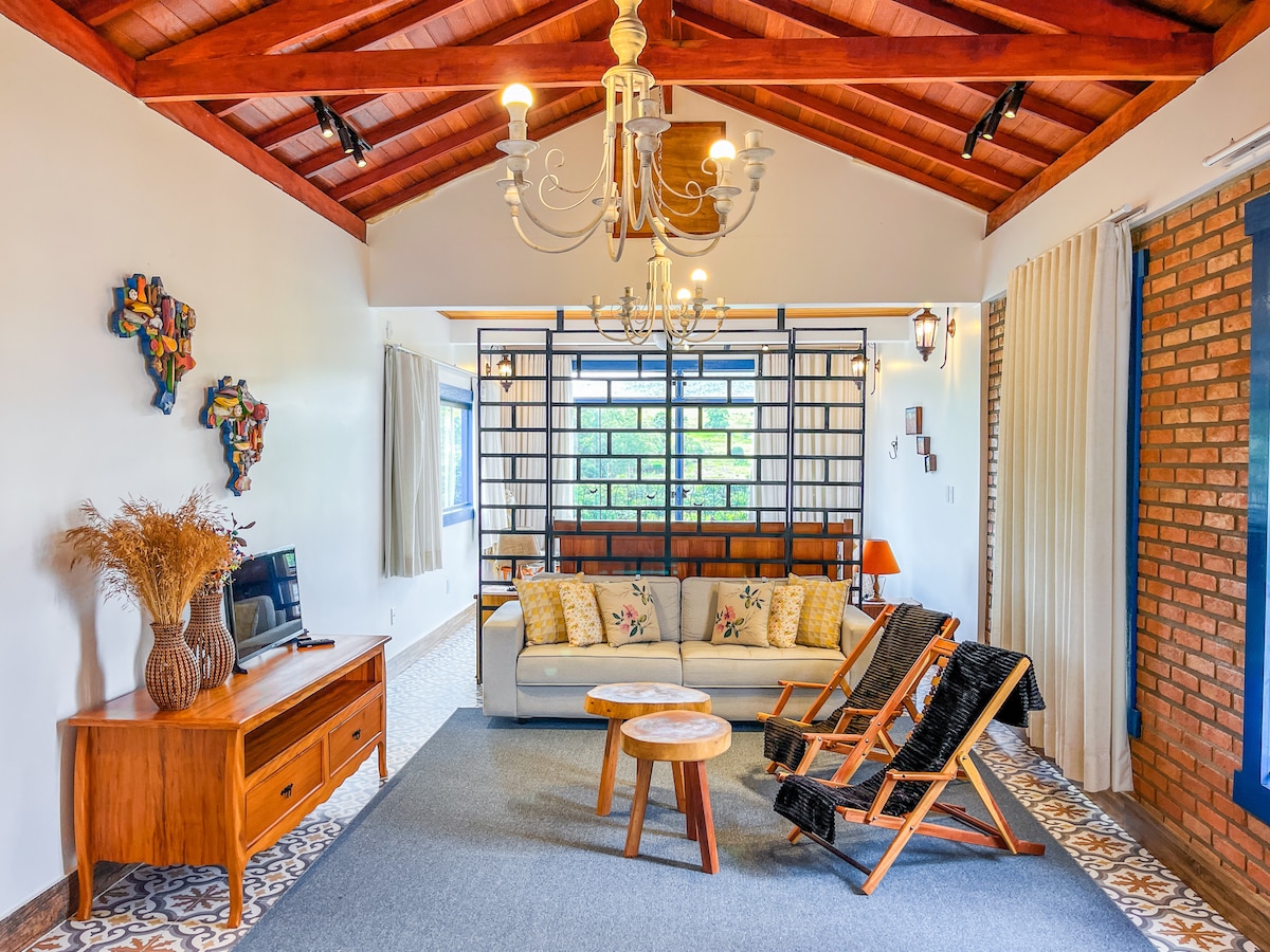 A spacious living area is featured, characterized by a wooden ceiling and two large windows that offer natural light. Comfortable seating is arranged around a small wooden table. Decorative elements include wall art and a console table with a television, enhancing the room's inviting ambiance.