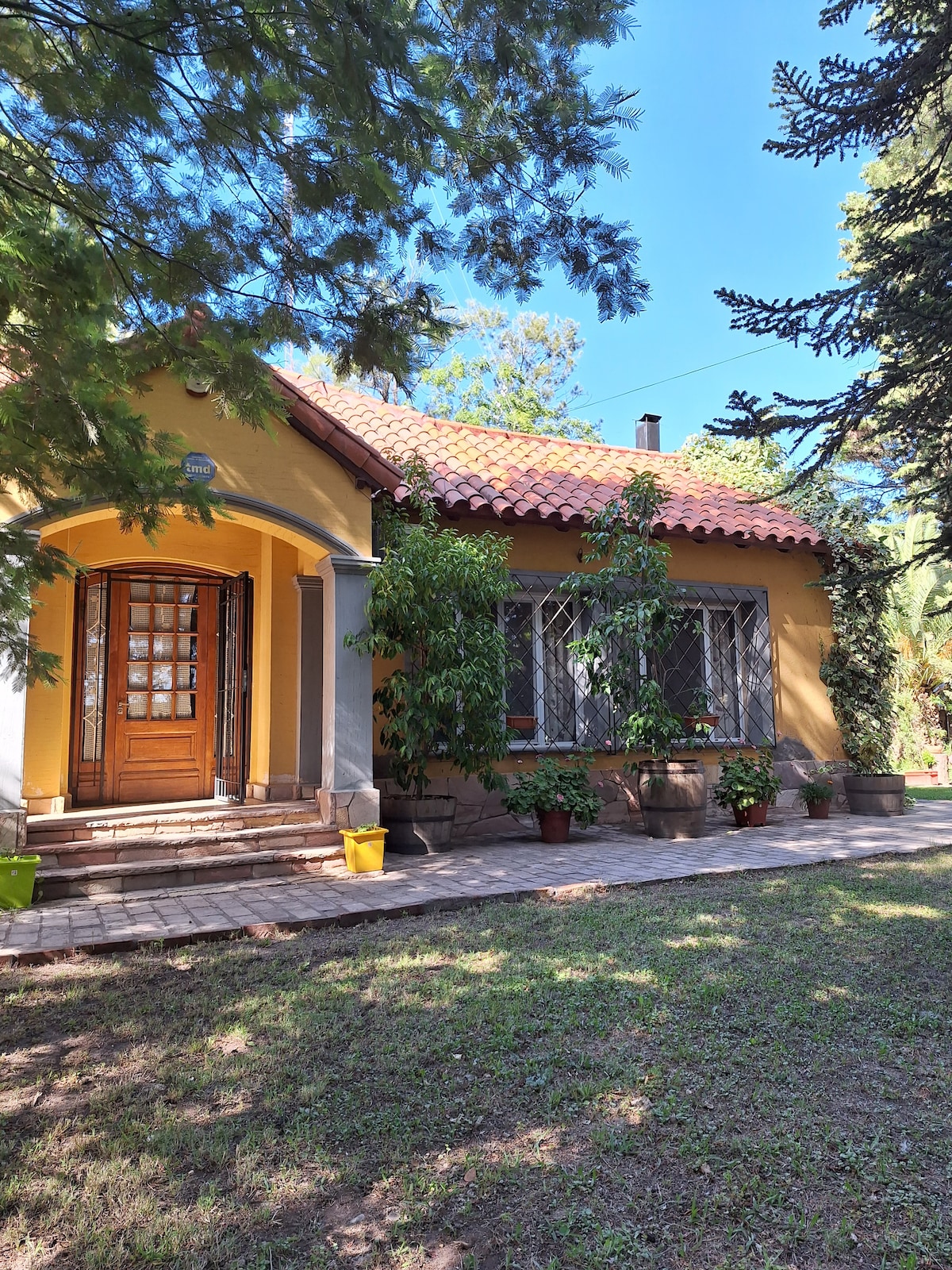 A charming yellow house features a welcoming entrance with a wooden door and tiled roof, framed by greenery. Potted plants are arranged along the facade, while a stone pathway leads to the door. Sunlight filters through the surrounding trees, highlighting the inviting exterior.