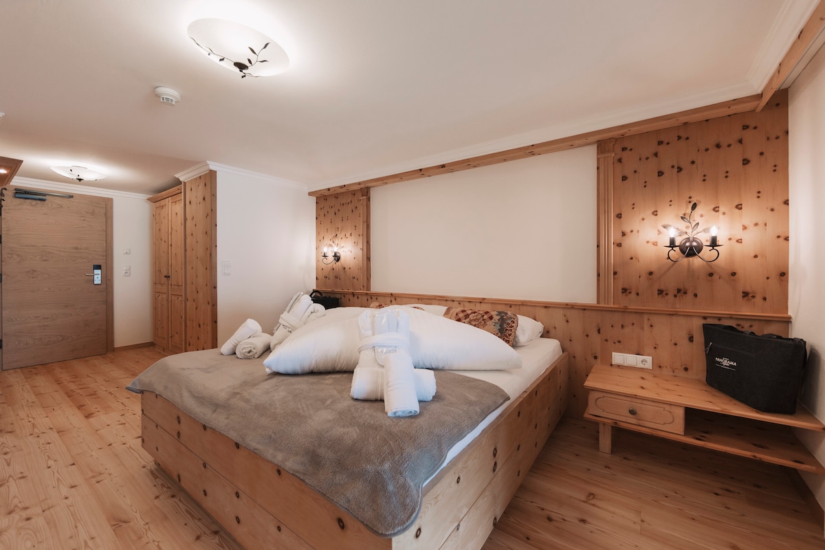 A wooden-paneled room features a spacious bed topped with fluffy pillows and a cozy blanket. Soft light illuminates the room from wall sconces on either side of the bed. Natural wooden flooring adds warmth to the atmosphere, complemented by a side table equipped with a telephone.