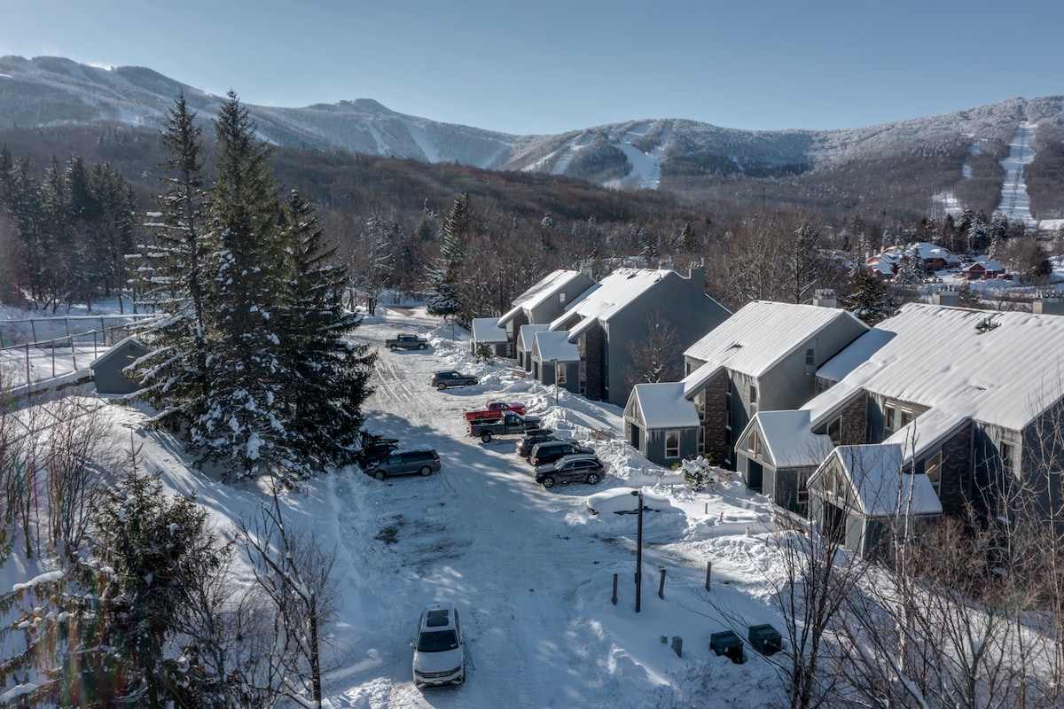 An aerial view captures the snow-covered Whiffletree condo complex surrounded by trees. Several parked vehicles are seen in the snow-covered parking area, with distant mountains visible in the background, offering a serene alpine setting.