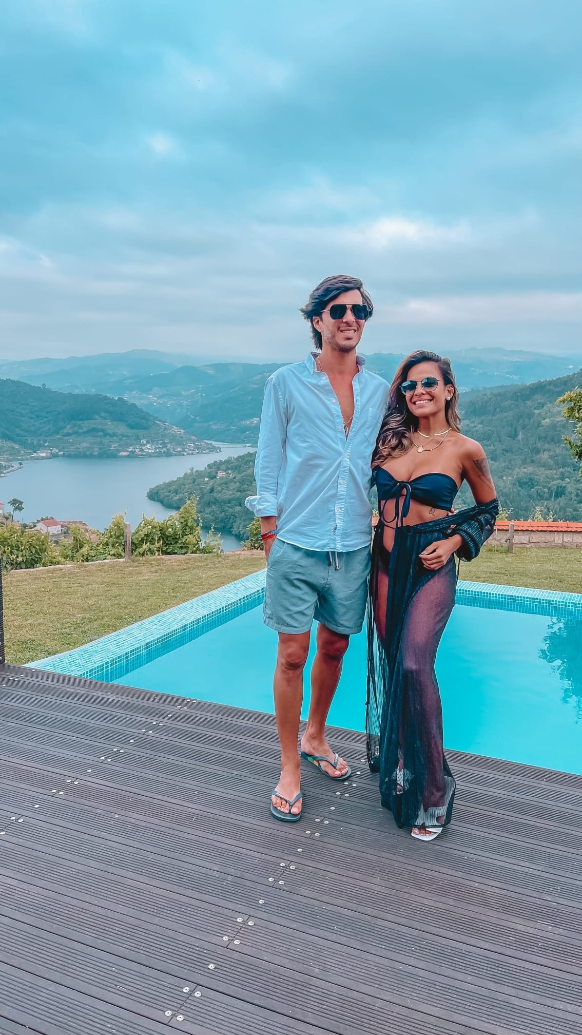 The image captures two individuals standing by a swimming pool, with a scenic view of rolling hills and a river in the background. The pool reflects the cloudy sky, while wooden decking surrounds it. Green landscapes and distant hills add to the tranquil atmosphere.