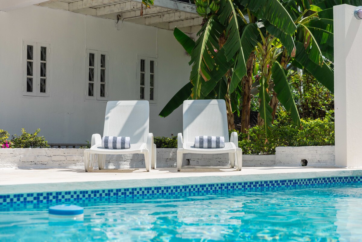Two white lounge chairs are positioned beside a clear blue pool, with beach towels neatly arranged on each. Lush greenery and palm trees frame the scene, while sunlight filters through the surrounding vegetation, creating a tranquil outdoor space.