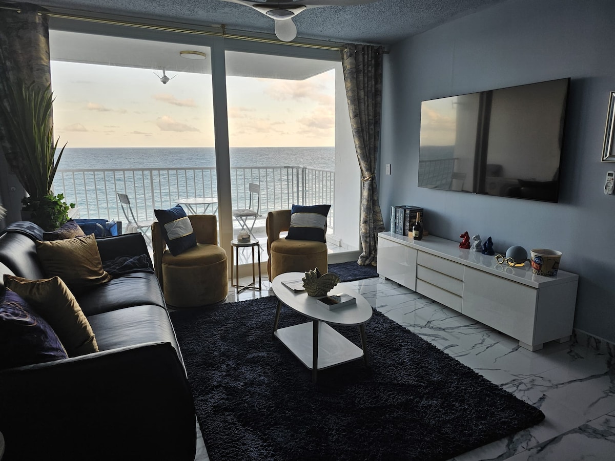 A comfortable living area features a dark rug, a sleek white coffee table, and a television mounted on the wall. Two armchairs and a sofa offer seating, while large windows provide a view of the ocean and natural light. A small dining area is visible on the balcony.