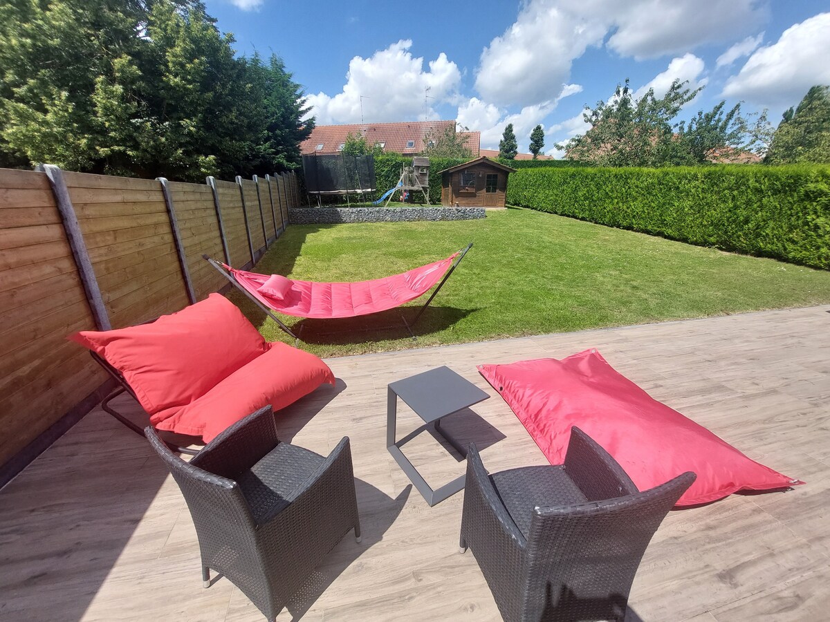 An outdoor space is furnished with two black wicker chairs and a small table. Bright red lounge cushions and a hammock add comfort to the grassy area, which is bordered by tall green hedges. A swing set and a wooden shed are visible in the distance.