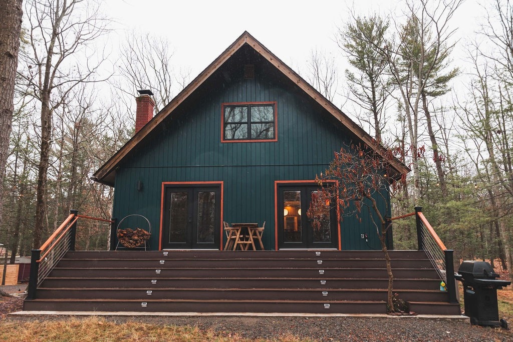 The modern chalet features a striking blue exterior with orange accents and large windows. A spacious deck with stepped entrances leads to a welcoming entry. Surrounded by trees, the setting offers a serene outdoor atmosphere, perfect for relaxation.