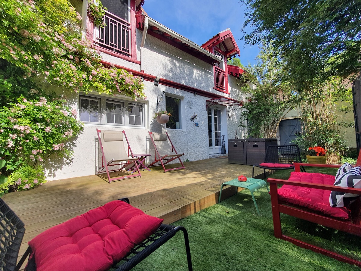 The outdoor terrace is surrounded by lush greenery, featuring wooden decking and vibrant red outdoor furniture. Two reclining chairs and a seating area invite relaxation, with small planters and colorful flowers adding a touch of nature. A shed is visible in the background.