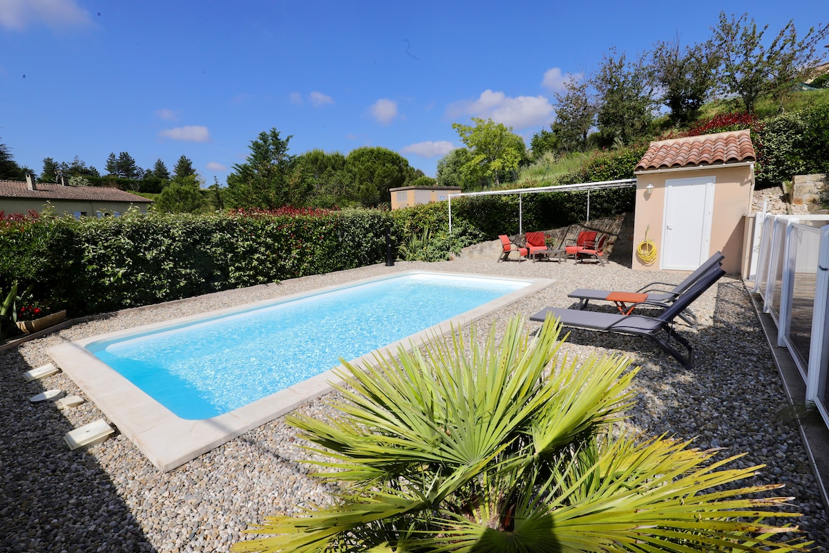 A rectangular swimming pool is surrounded by a landscaped area featuring gravel flooring and lush green shrubs. Sun loungers are positioned nearby, and a small shed is present in the background, integrated into a serene garden setting.