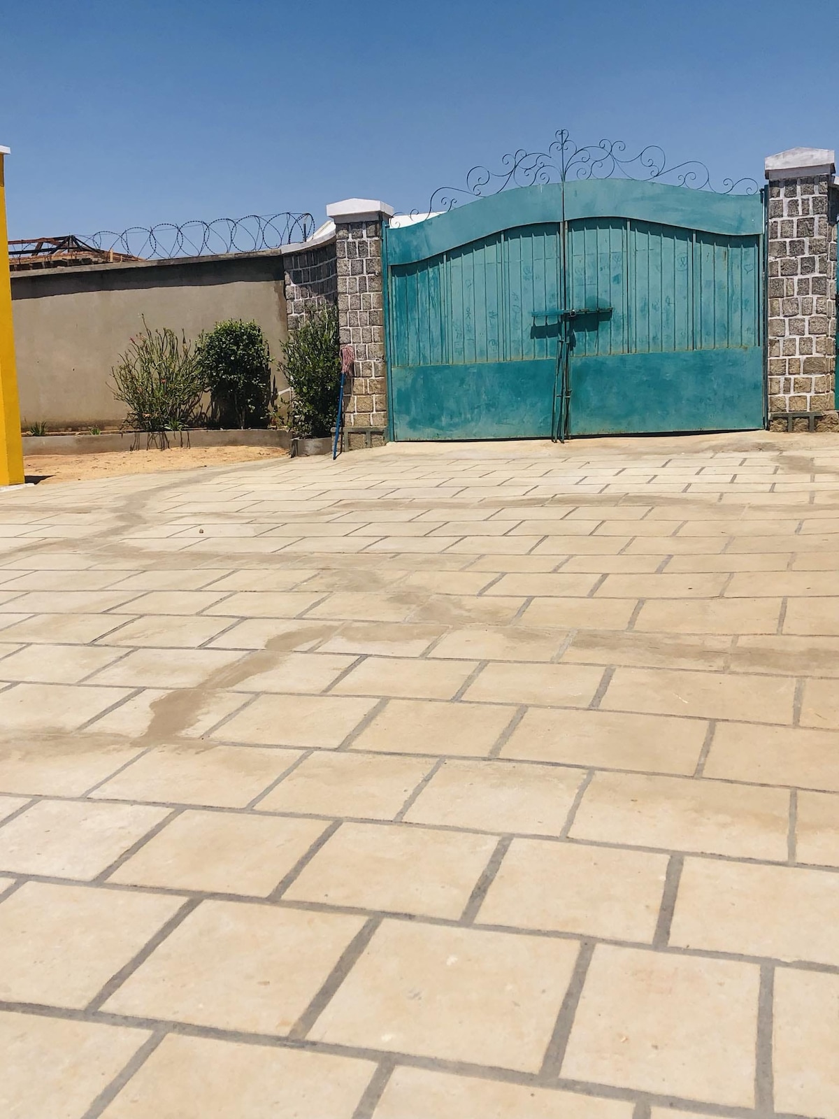 A spacious entrance is shown, featuring a large turquoise gate framed by stone walls. The surrounding area includes neatly arranged stone pavement and scattered greenery, contributing to a welcoming arrival space.