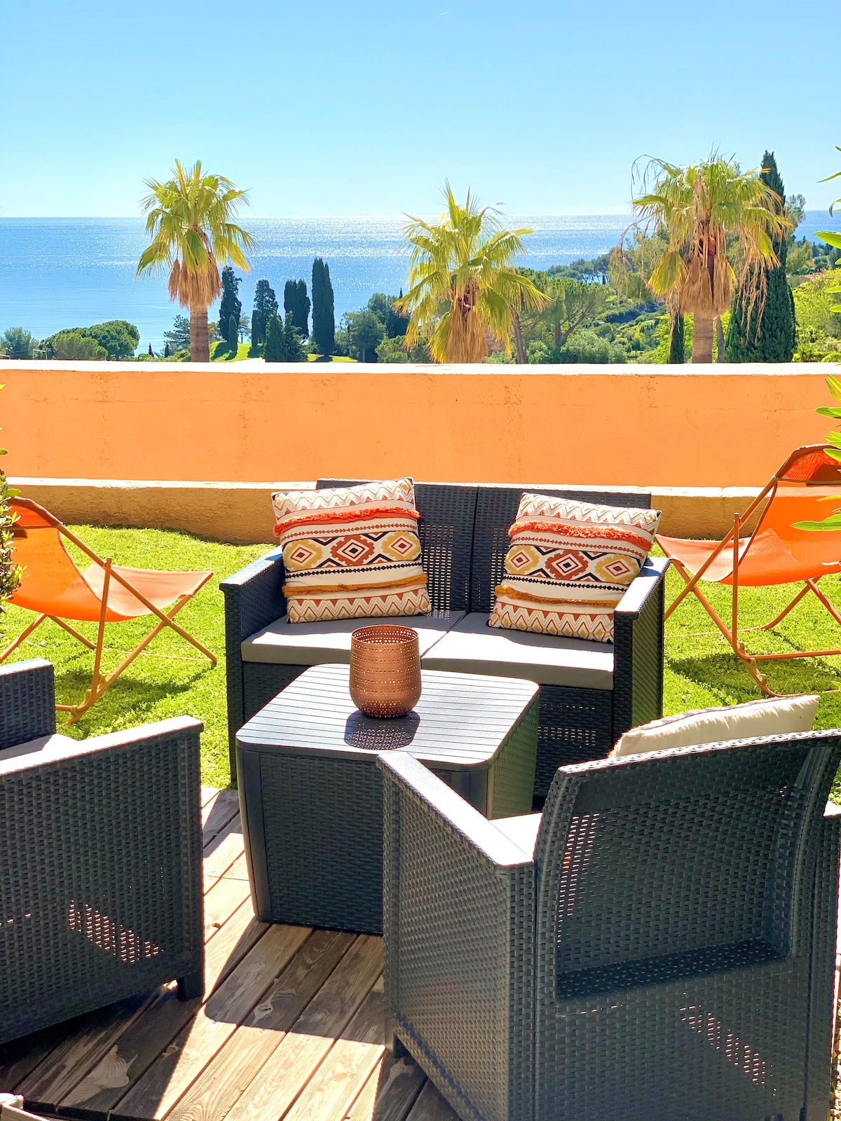 An outdoor seating area is displayed, featuring four cushioned chairs arranged around a small table. The backdrop consists of a lush garden with palm trees and a distant view of the sea. Bright sunlight enhances the tranquil setting, creating a relaxing atmosphere.