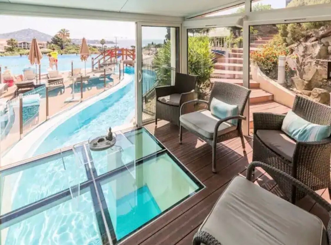 A modern seating area is presented with comfortable wicker chairs overlooking a swimming pool. Large glass panels provide views of the pool and outdoor space, allowing natural light to fill the room. A glass-top table adds a distinctive element to the seating area.