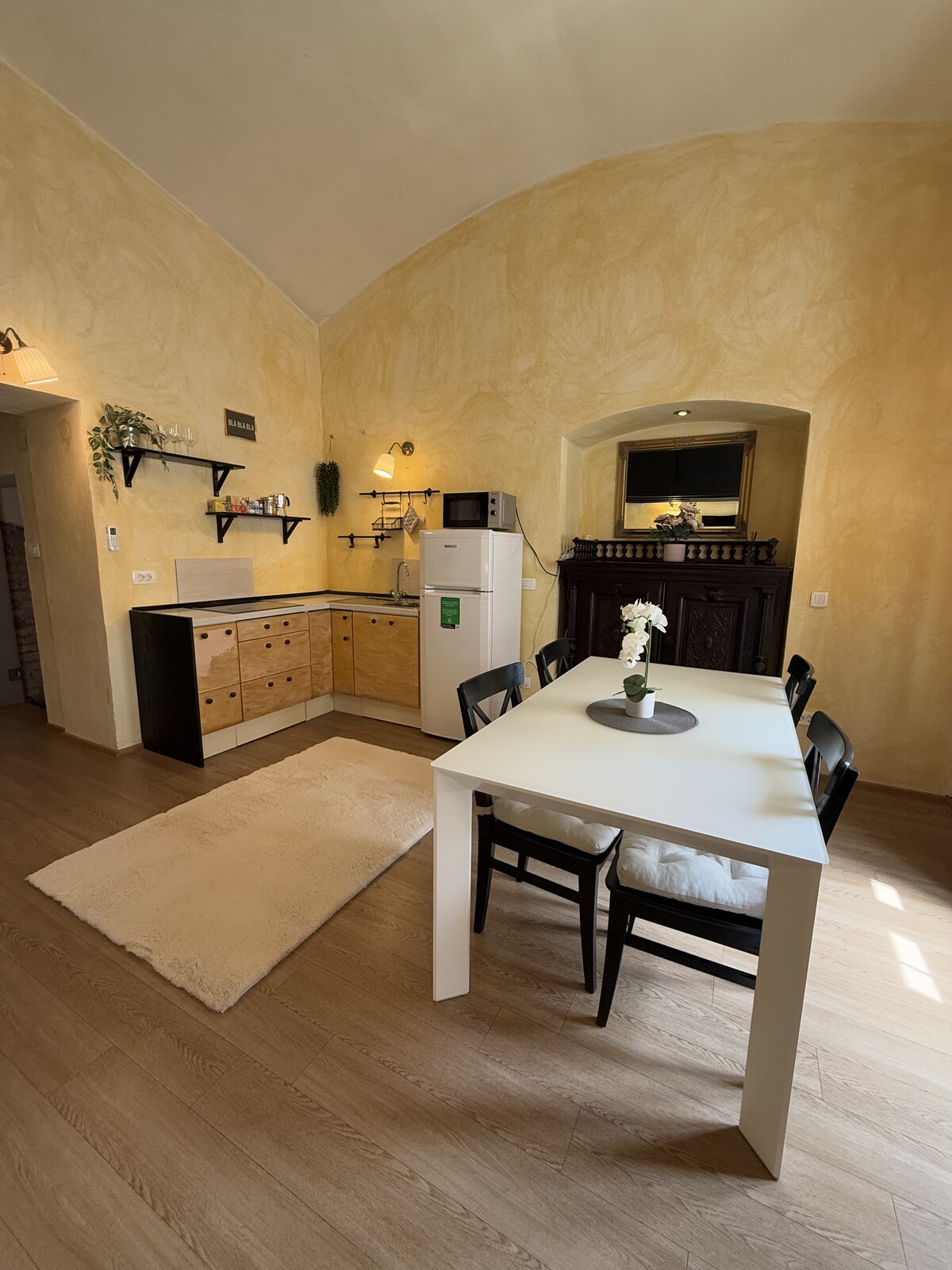 The kitchen area features a spacious layout with wooden cabinetry and a white dining table. Black chairs surround the table, while a small vase with a flower adds a touch of decor. Natural light enhances the warm, yellow walls, creating a welcoming environment.