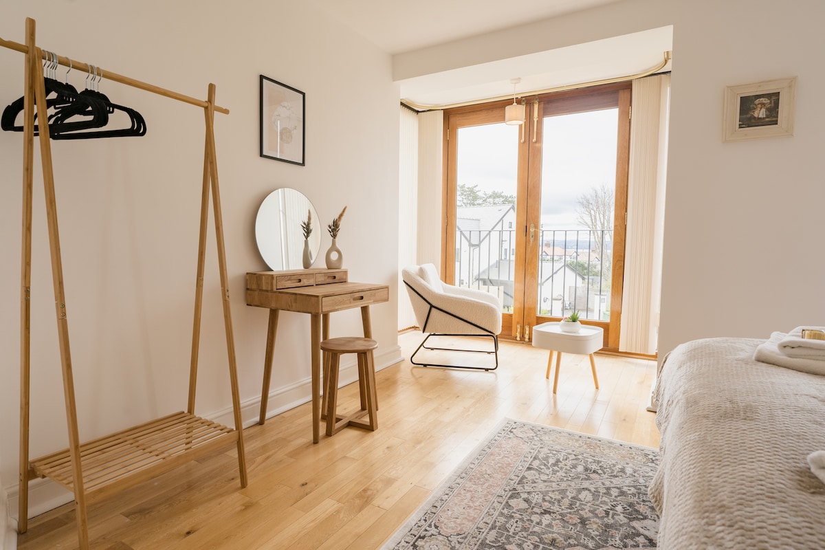 A bright and airy room features a comfortable chair and a small side table next to large glass doors that open to a balcony. A wooden vanity with a mirror and a stool is positioned beside a clothing rack, with soft light enhancing the natural wood tones.
