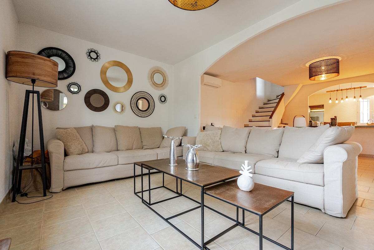 A spacious living area is presented, featuring a light-colored sectional sofa arranged around a modern coffee table. Various decorative mirrors adorn the wall, creating visual interest. In the background, a staircase leads to an upper level, while soft lighting fixtures hang above.