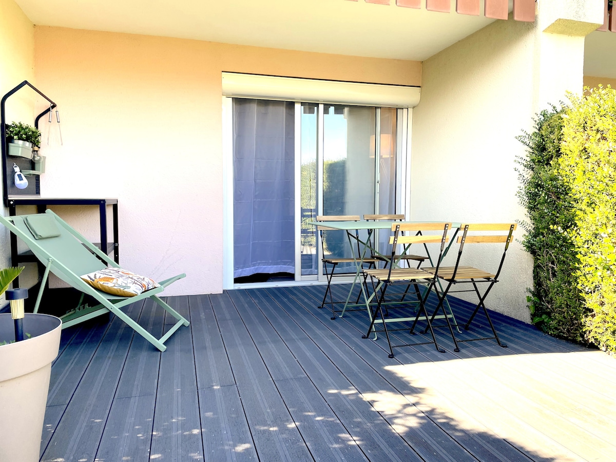 A spacious terrace features a light green lounge chair and a small dining table surrounded by four folding chairs. Large sliding doors lead into the interior space, with a view of greenery just beyond the railing.