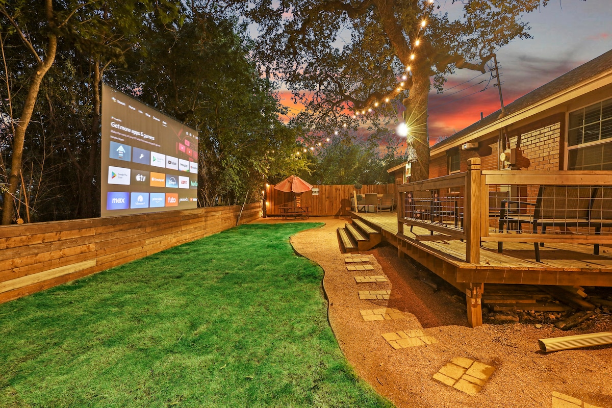 A spacious outdoor area is shown, featuring a large screen set against a backdrop of trees. Soft lights are strung above, illuminating the space. A deck with seating is visible, along with a pathway made of stone leading through the manicured grass.