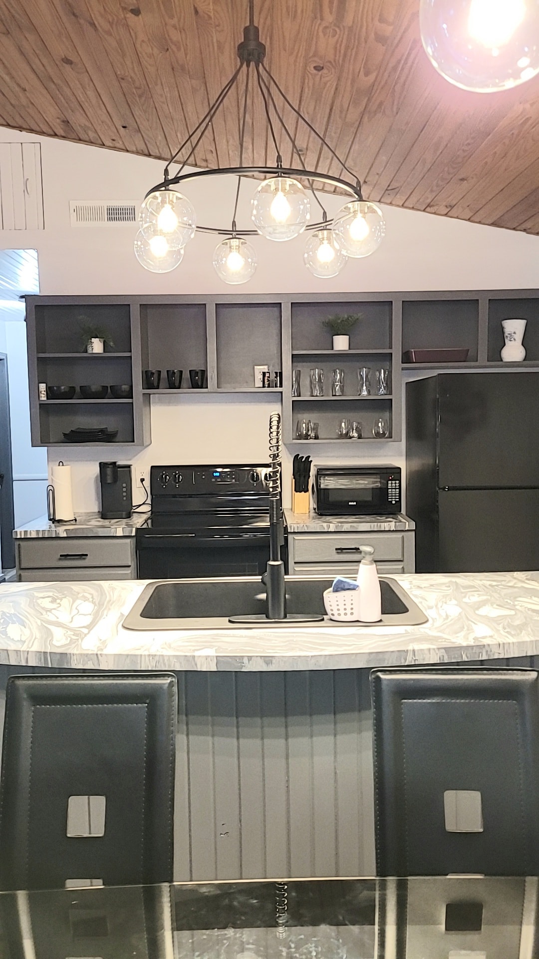 The kitchen features modern black appliances and sleek grey cabinetry. A spacious island with a marble countertop serves as an area for preparation and dining. Pendant lighting hangs above, providing illumination to the workspace, while various kitchenware can be seen on open shelves.
