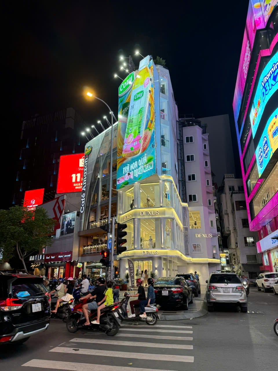 A modern building stands prominently in a busy urban setting at night, with bright advertising displays illuminating the façade. Vehicles are seen moving along the road, while pedestrians navigate the crosswalk. The vibrant atmosphere reflects the heartbeat of the city.