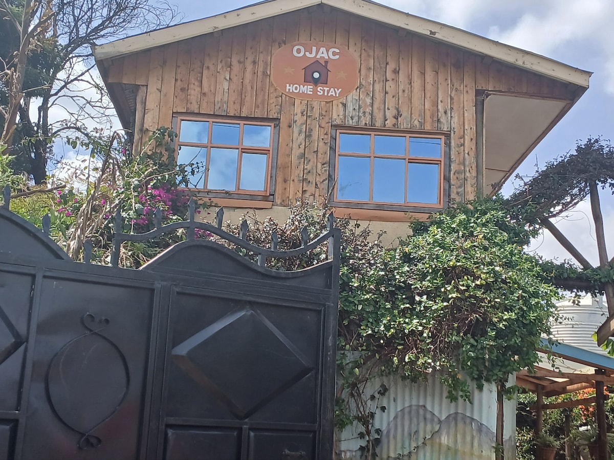 The exterior of Ojac Home Stay is displayed, featuring a wooden structure with multiple windows framed in dark wood. The entrance is marked by a welcoming sign. Lush greenery and flowering plants surround the building, contributing to a natural, inviting environment.
