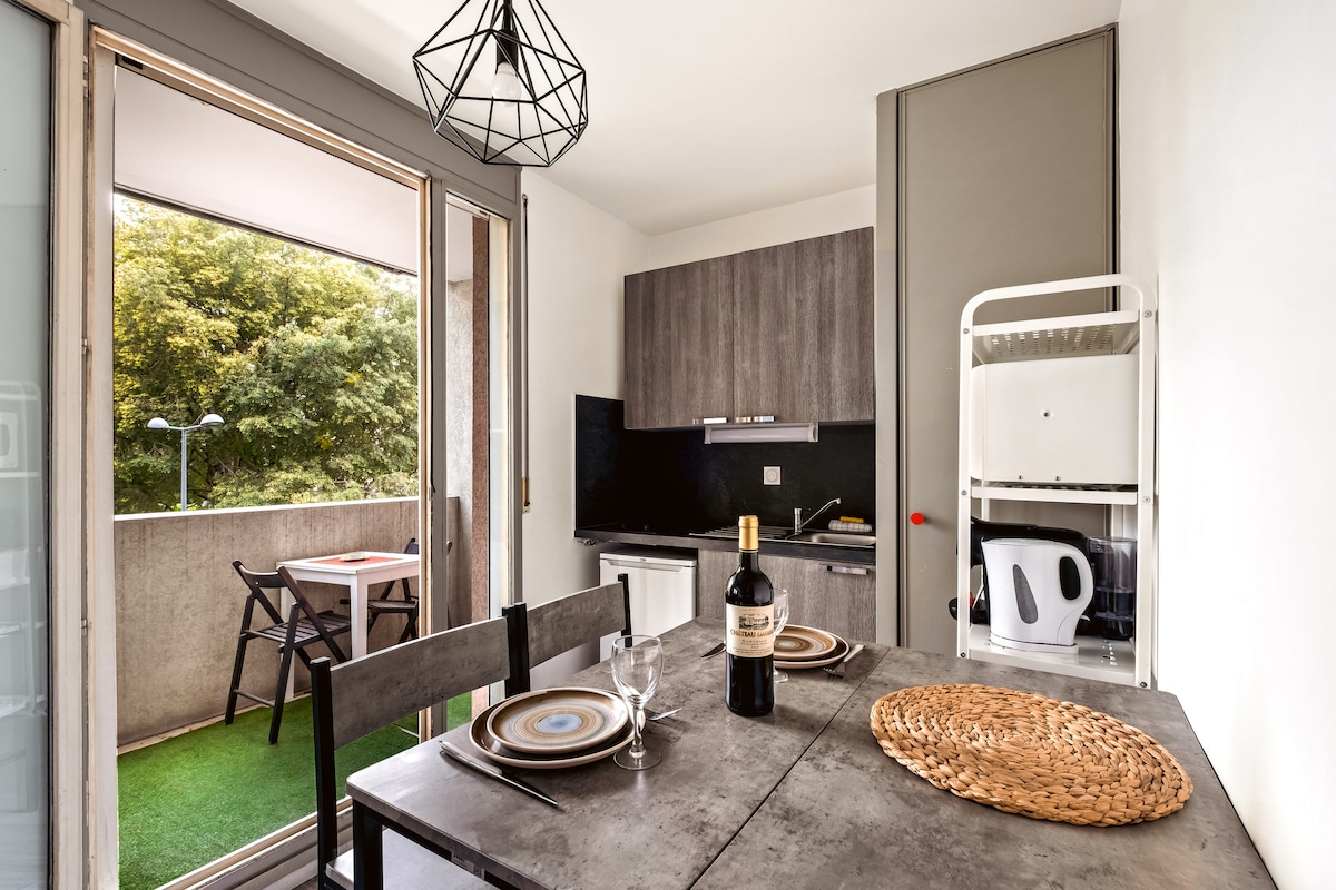 An inviting dining area is captured, featuring a modern table set for two. A bottle of wine sits centrally on the table, flanked by plates and cutlery. A kitchenette is visible, equipped with essential appliances, and sliding glass doors open to a small balcony with outdoor seating.