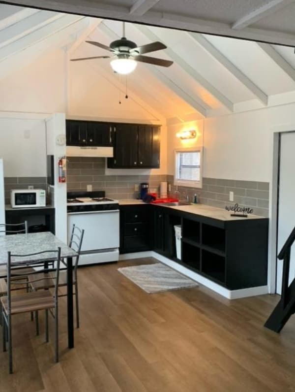 A compact kitchen area is featured, equipped with a stove, microwave, and refrigerator, complemented by dark cabinetry and a bright backsplash. A small dining table with chairs is visible, and natural light enters through the window, highlighting the efficiency of the space.