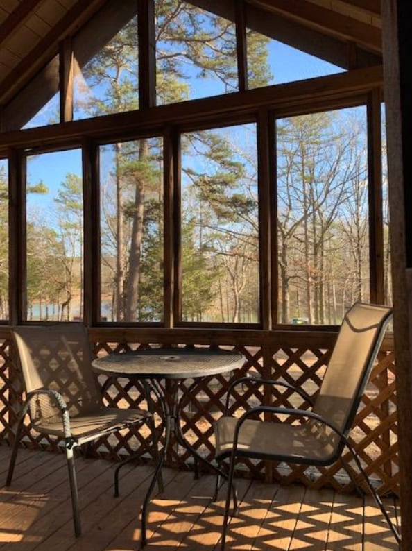 A screened-in porch is visible, featuring a round table and two chairs made of metal. Natural light filters through the screens, highlighting the wooden flooring. Outside, trees and a glimpse of water can be seen, creating a serene outdoor atmosphere.