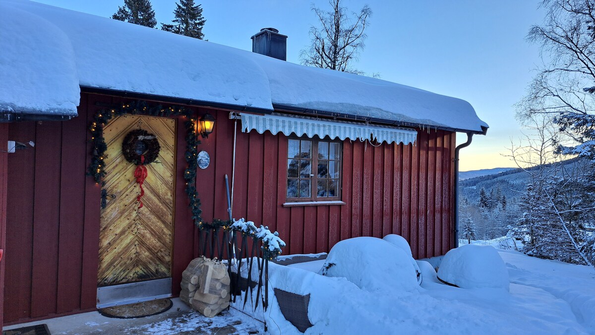 The exterior of a red wooden cabin is shown, dusted with fresh snow. A warmly lit entrance features a decorative wreath and wooden door with a chevron pattern. Snow-covered furniture and a serene winter landscape are visible in the background.