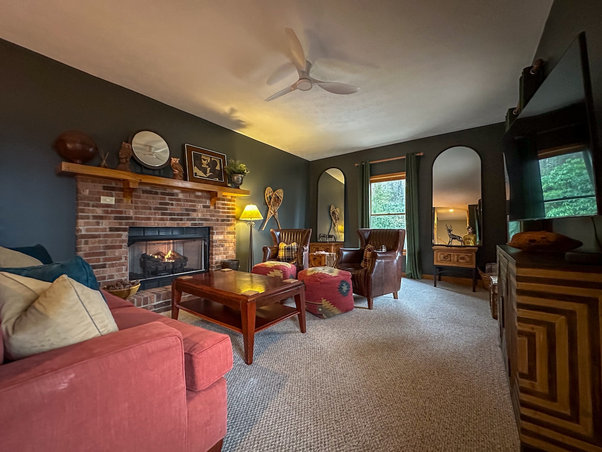 A spacious living room features a cozy fireplace surrounded by tasteful decor. Comfortable seating arrangements include two upholstered armchairs and a pink sofa. Large windows provide natural light and views of the outdoors, while a tabletop centerpiece adds to the room's inviting feel.