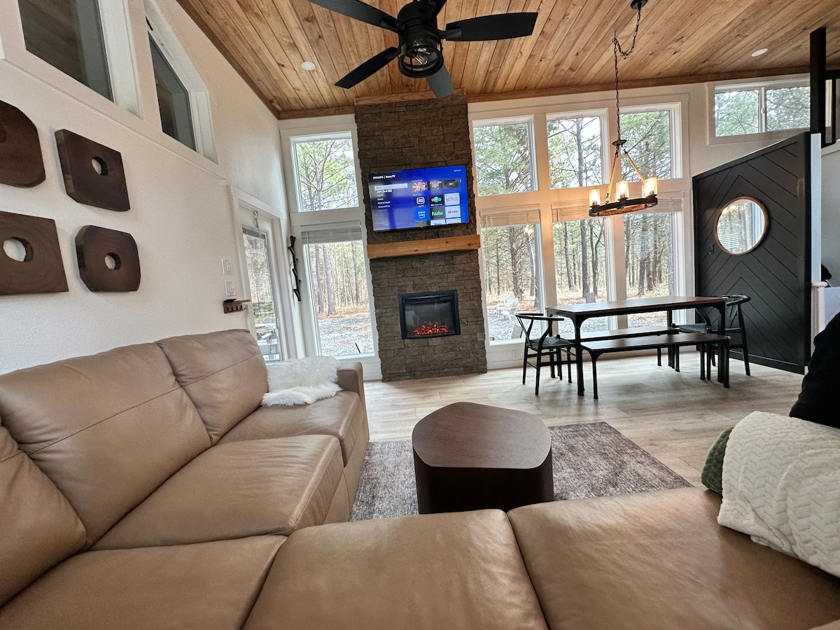 A welcoming living area is accentuated by large windows, allowing natural light to fill the space. A sleek brown leather sofa is positioned for comfort while a modern fireplace provides warmth. A dining table sits nearby, complementing the rustic design of the wooden ceiling.