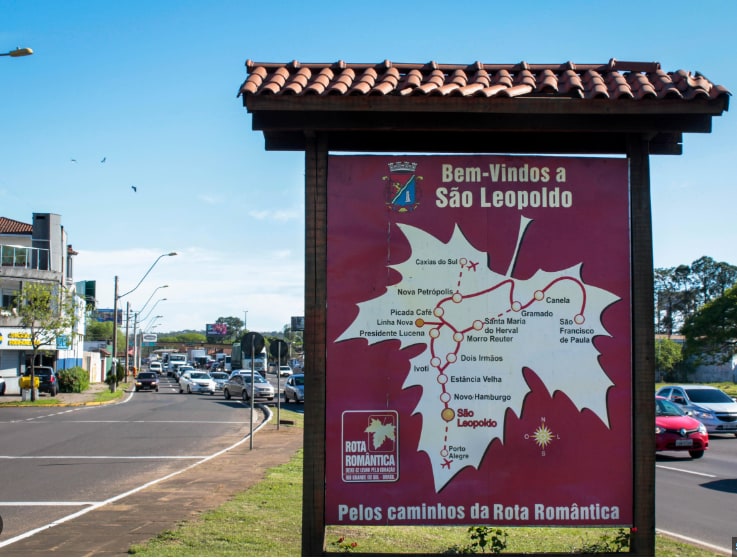 A welcome sign for São Leopoldo is prominently displayed, outlining nearby locations on a map. The background features a bustling street with trees and buildings visible. The clear blue sky adds brightness to the scene, enhancing the friendliness of the area.