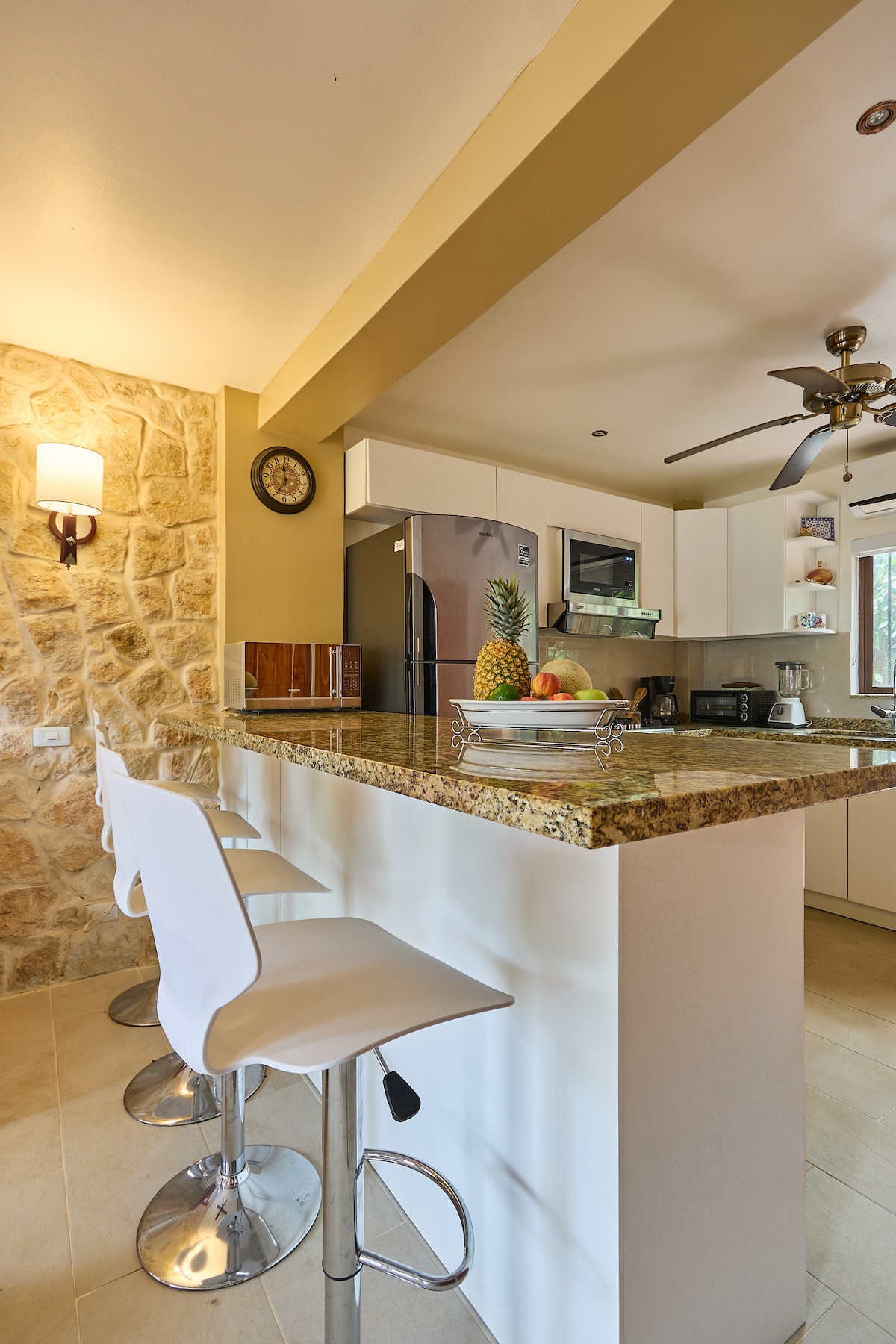 A modern kitchen area features a granite countertop with three high-backed bar stools. Natural light illuminates the space, highlighting the stone accent wall and contemporary cabinetry. Kitchen appliances and decorative fruit enhance the inviting atmosphere of the culinary space.