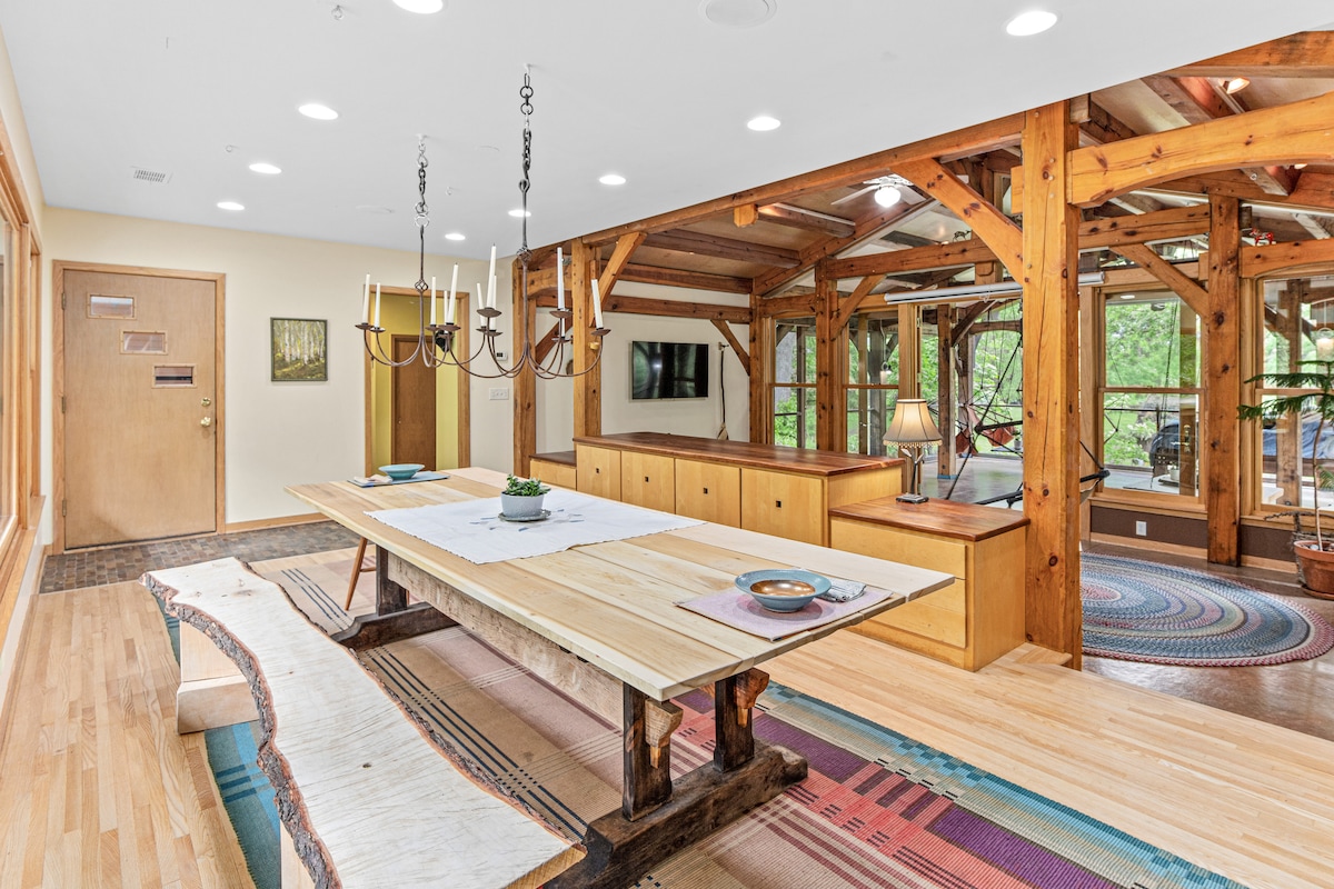 A spacious dining area features a large wooden table, complemented by a unique base. Surrounding the table are simple plates and a small potted plant. Natural wood beams and large windows offer a view of greenery, creating an open and bright environment.
